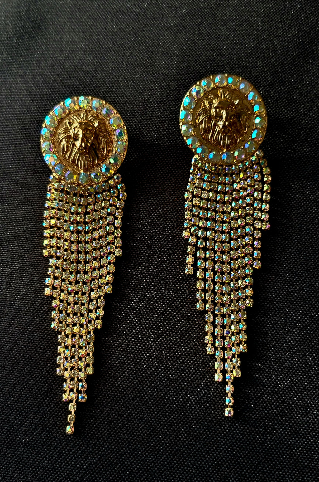 Pair of gold earrings with lion emblem and dangling rhinestone tassels on a black background